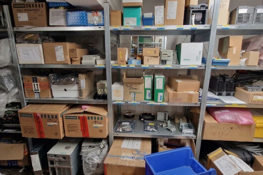 Stellingen met reservedelen: kartonnen dozen, elektronicamodules en antistatische zakken, met gelabelde schapranden
